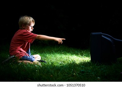 Child Blond Boy Watching TV Horror / Thriller. Outside In The Garden Sitting On Grass Lawn At Night. Concept Of Inappropriate Movies For Children