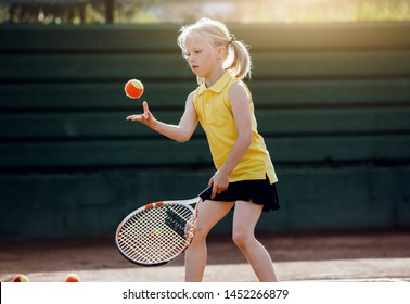 A child blond girl in a yellow polo playing tennis at the court on a sunset