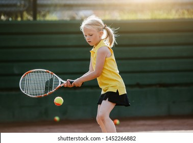 A child blond girl in a yellow polo playing tennis at the court on a sunset - Powered by Shutterstock