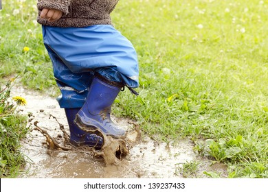 Child In Boots Jumping In Muddy Puddle After Rain.