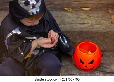 Child Boy In Black Halloween Costume Unwrapping Candy Sitting On Steps Near Orange Pumpkin Bucket