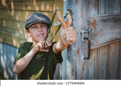 child boy in a cap takes aim with a slingshot to shoot at a target. Play around as a child in the village on vacation. Carefree childhood mischief maker.