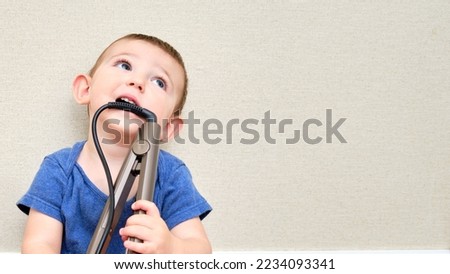 Child boy chewing on the electric wire of a curling iron, copy space. Toddler baby plays with hot curling tongs. Kid age one year