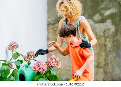 Child Boy With Down Syndrome In The Garden And Learning To Plant Flowers With His Blond Mother Gardener In Black Glowes. Affection Female With Her Son Working In Garden Outdoor Shot.