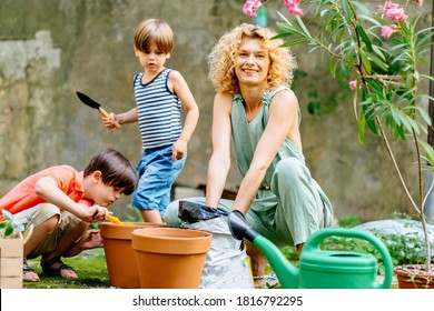 Child Boy With Down Syndrome In The Garden And Learning To Plant Flowers In Pot With His Blond Mother Gardener In Black Gloves Family Having Fun Outdoor. Developmental Activities For Children Concept.