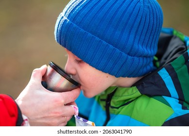 A child boy drinks warm fresh tea from a thermos cup, quenching his thirst after active play while walking outdoors. Give me a drink. Picnic water.