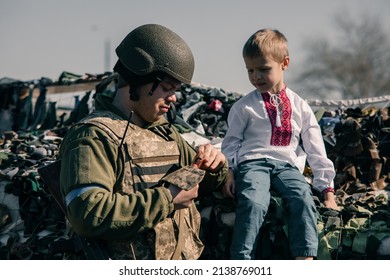 Child boy in embroidered shirt sits on sandbags in checkpoint near Ukrainian territorial defense warrior. Concept of russian military invasion in Ukraine. War in Ukraine and Europe. Children and war.