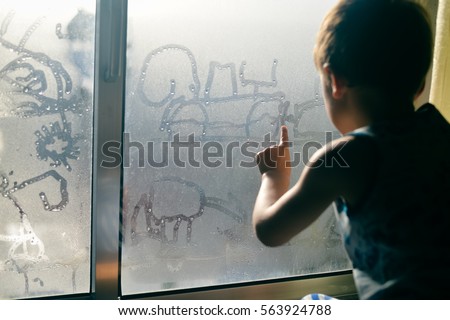 Child boy with hand draws on cold fogged window, closeup image. back view, blur sun light background