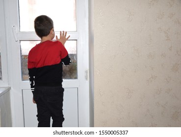A child, a boy at home left alone, looks out the window and waits for mom. A boy is standing at the window looking at the street and waiting for his mother.