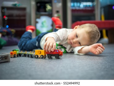 Child Boy Play With Wooden Train While Lying On Floor, Build Toy Railroad At Home Or Daycare. Toddler Boy Play With Crane And Cars Educational Toys For Preschool, Kindergarten Child. Indoor Playground