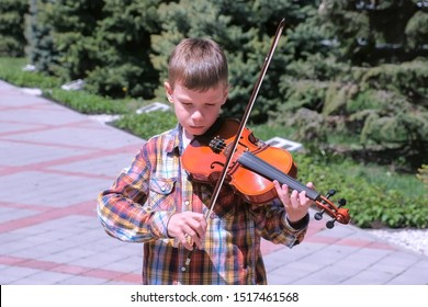 Child Boy Is Playing Violin Standing In Alley On Street In City. Portrait Of Little Musician With Instrument. Education Learn Play Violin Boy Teen Violinist Instrumentalist Performance Melody.