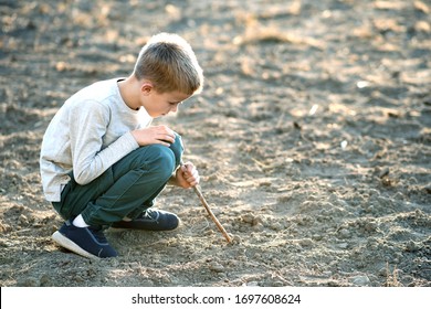 Child Boy Playing With Wooden Stick Digging In Black Dirt Ground Outdoors.