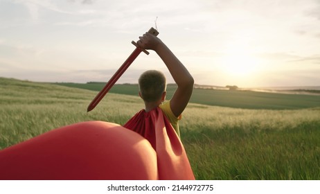 Child boy plays superhero. Child Game. Boy in red cloak stands with sword raised in his hand up sword, on field, depicting medieval knight. Child waves toy sword, childhood dreams. Imagination