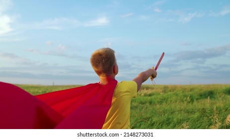 Child boy plays superhero. Child Game, Imagination. Boy, child in red cloak go with sword raised in his hand up sword, on field, depicting medieval knight. Child waves toy sword, childhood dreams.