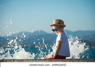 Child Boy Sitting On The Beach With Water Splash. Happy Boy In Hat And Sunglasses On Summer Holiday Vacation. 