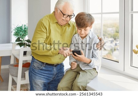 Child boy using mobile phone together with senior elderly grandfather sitting in living room at home. Grandpa browsing internet on smartphone with grandchild. Family technology and leisure concept.
