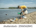 A child boy volunteer in gloves tidying up rubbish plastic garbage from the riverbank. Children take care of nature.