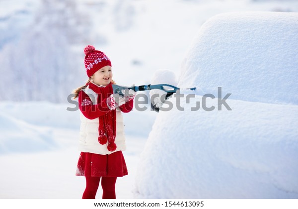 Child brushing snow off car after storm. Kid with\
winter brush and scraper clearing family car after overnight snow\
blizzard. Family Christmas vacation in the mountains. Kids\
shoveling snow.