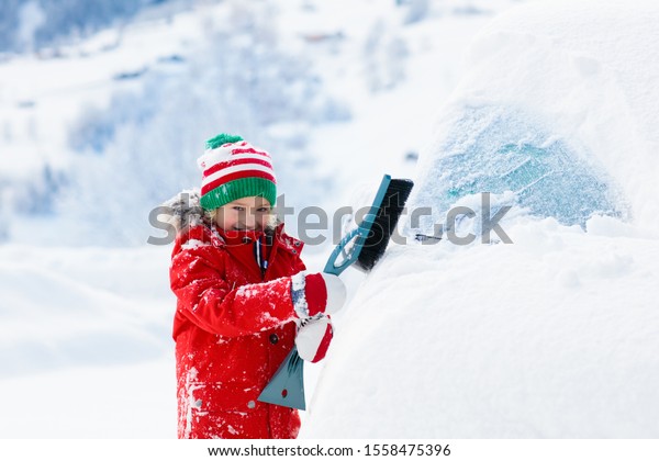 Child brushing snow off car after storm. Kid with\
winter brush and scraper clearing family car after overnight snow\
blizzard. Family Christmas vacation in the mountains. Kids\
shoveling snow.