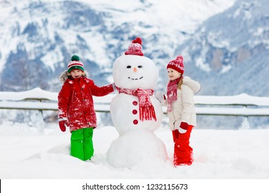 Child Building Snowman. Kids Build Snow Man. Boy And Girl Playing Outdoors On Snowy Winter Day. Outdoor Family Fun On Christmas Vacation In The Mountains. Children Play In Swiss Mountain Landscape.