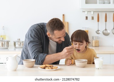 Child care concept. Young loving single father feeding his little son with cereals at home, eating breakfast together at kitchen in morning, empty space