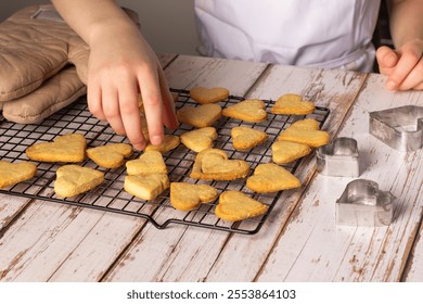 A child carefully places freshly baked heart-shaped cookies onto a cooling rack. The setting features a rustic wooden table and metal cookie cutters in the background, capturing a warm baking moment. - Powered by Shutterstock