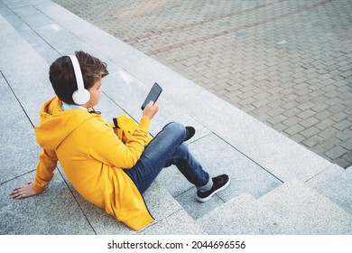 Child Caucasian Teen Boy In Yellow Hoodie With Wireless Headphones Sitting Outdoors Using Smartphone. Communication On The Go. Candid Blogging Vlogging Surfing. Music. Video Call.