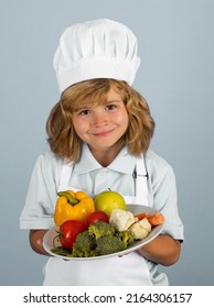 Child Chef Cook Hold Plate With Vegetables Prepares Food On Isolated Grey Studio Background. Kids Cooking. Teen Boy With Apron And Chef Hat Preparing A Healthy Vegetables Meal In The Kitchen.