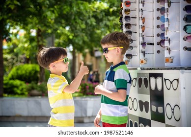 A child and a choice of sunglasses. Two little boys are standing in sun-proof glasses against the background of a shop window with glasses. Kid help choose each other.