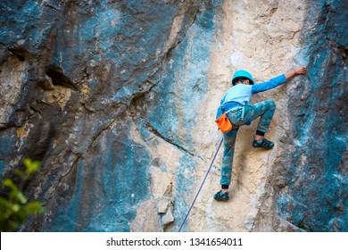 A Child Climber Climbs On A Rock. The Boy In The Helmet Climbs Up The Cliff. Strong Kid Overcomes A Difficult Climbing Route On A Natural Relief. Extreme Sport. Insurance And Safety.