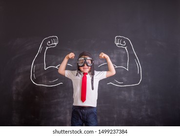 Child With Confidence Ready To Defense From Bullying. School Boy In Uniform And Pilot Helmet And Glasses. Kid Showing Bicep Muscles With Muscles Drawn On Chalkboard. Back To School