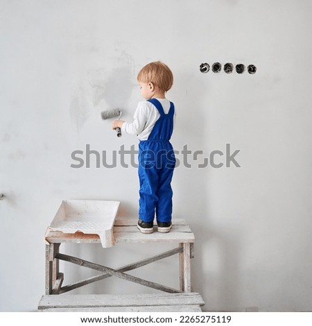 Child construction worker standing on wooden table and painting white wall in apartment. Kid in work overalls using paint roller while playing at home under renovation.