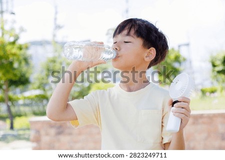 Child cools down with a portable handheld fan while drinking water outdoors during hot summer weather. Hydration and personal cooling help stay comfortable during extreme heat and sunny conditions.