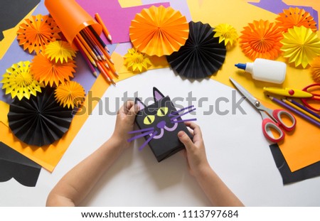 The child creates a gift box of a black cat. A party for Halloween. Children's hands make a master class. Craft for kids. Materials for creativity of orange, purple and black colors. School, education