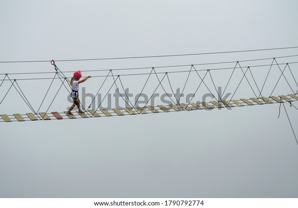A child crosses\
an abyss in the fog on a rope bridge with fall protection.\
Attraction in the mountains.