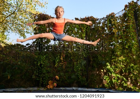 Child cute blond girl playing and jumping on trampoline with greenery background