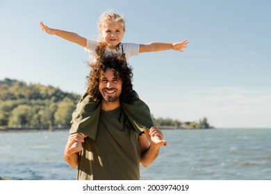 Child daughter sits on her dad shoulders outdoors on a summer beach and looking at camera. Loving father holding little kid child piggyback on weekend walking along lake river