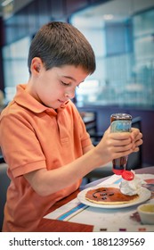 Child Decorates A Pancake With Chocolate Syrup Drawing A Face With A Smile