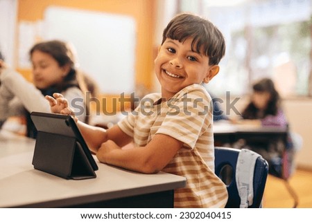 Child development in action as young kid learns to code with a tablet in school. Happy boy looking at the camera while sitting at his desk in a digital literacy class. Male student in primary school.