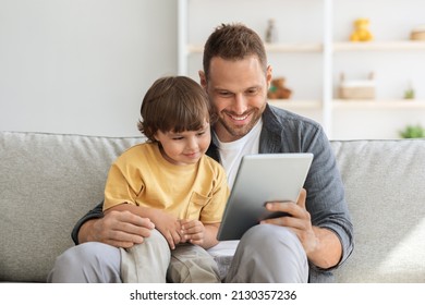 Child development content. Young positive father showing digital tablet to his cute little son, kid enjoying educational show online, sitting on sofa ta home - Powered by Shutterstock