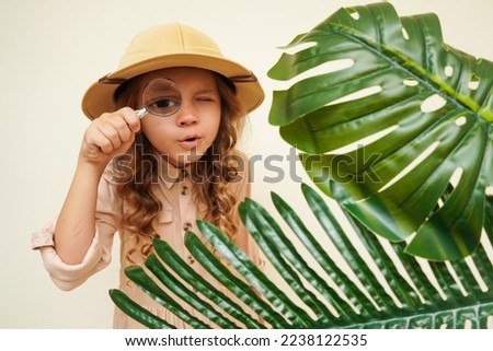 Child discoverer. Traveler child. Young explorer. A little girl in a safari hat with a magnifying glass looks carefully through the green palm leaves. Concept: search for adventure and treasure.