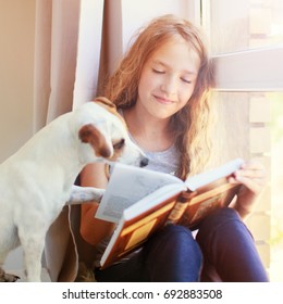 Child With Dog Reading Book At Home. Girl With Pet Sitting At Window At Read