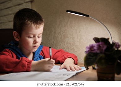 Child Is Doing Homework In The Light Of The Lamp In The Evening. A Boy Sits At A Table And Writes. Homework At His Desk