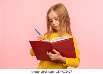 Child Doing Homework. Portrait Of Clever Little Girl Writing Poem In Book With Serious Concentrated Expression, Development Of Creative Child Abilities. Indoor Studio Shot Isolated On Pink Background