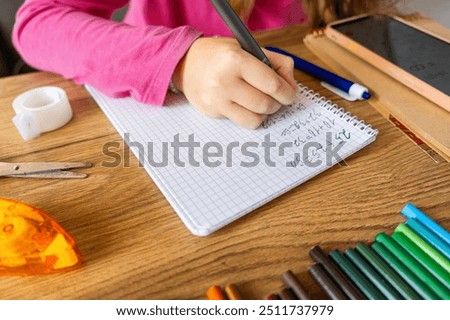Child doing math exercises in notebook using smartphone with stationery on wooden table. Girl hand writing mathematical equations as school homework. Learning, education using digital tools concept.