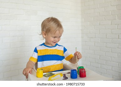 Child Draw With Paints, Playing At Home Sits At A Kid's Table. Todler With Educational Toys For Creativity. Boy Play At Home. Bright Paint Gouache Stained Face And Hands Of A Child. Kid Creative Plays