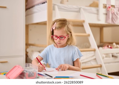 child drawing with colored pencils in cozy bedroom setting. young girl wearing red glasses, focused on art project at desk, surrounded by creative supplies in bright room