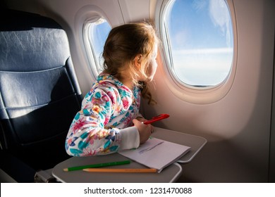 Child Drawing Picture With Crayons In Airplane. Little Girl Occupied While Flying In Aircraft. Travel With Family And Kids. Blue Sky And Sun Outside The Window