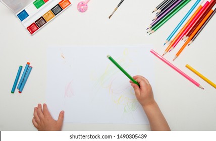 a child draws and crayons white lsita white table top view place copy child hand holds green pencil paint watercolor pen brush wax knives lie next