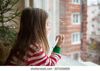 child draws snowflakes window and chalk marker  girl in New Year's pajamas windowsill against background live Christmas tree in pot in wicker basket 